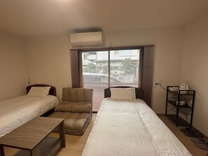 A hotel room with two beds, a chair, a table, a shelf with items, and a window with blinds.