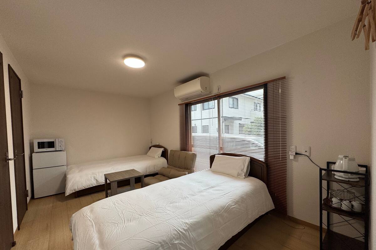 A small, clean room with two beds, a desk, a microwave, and a window with blinds.