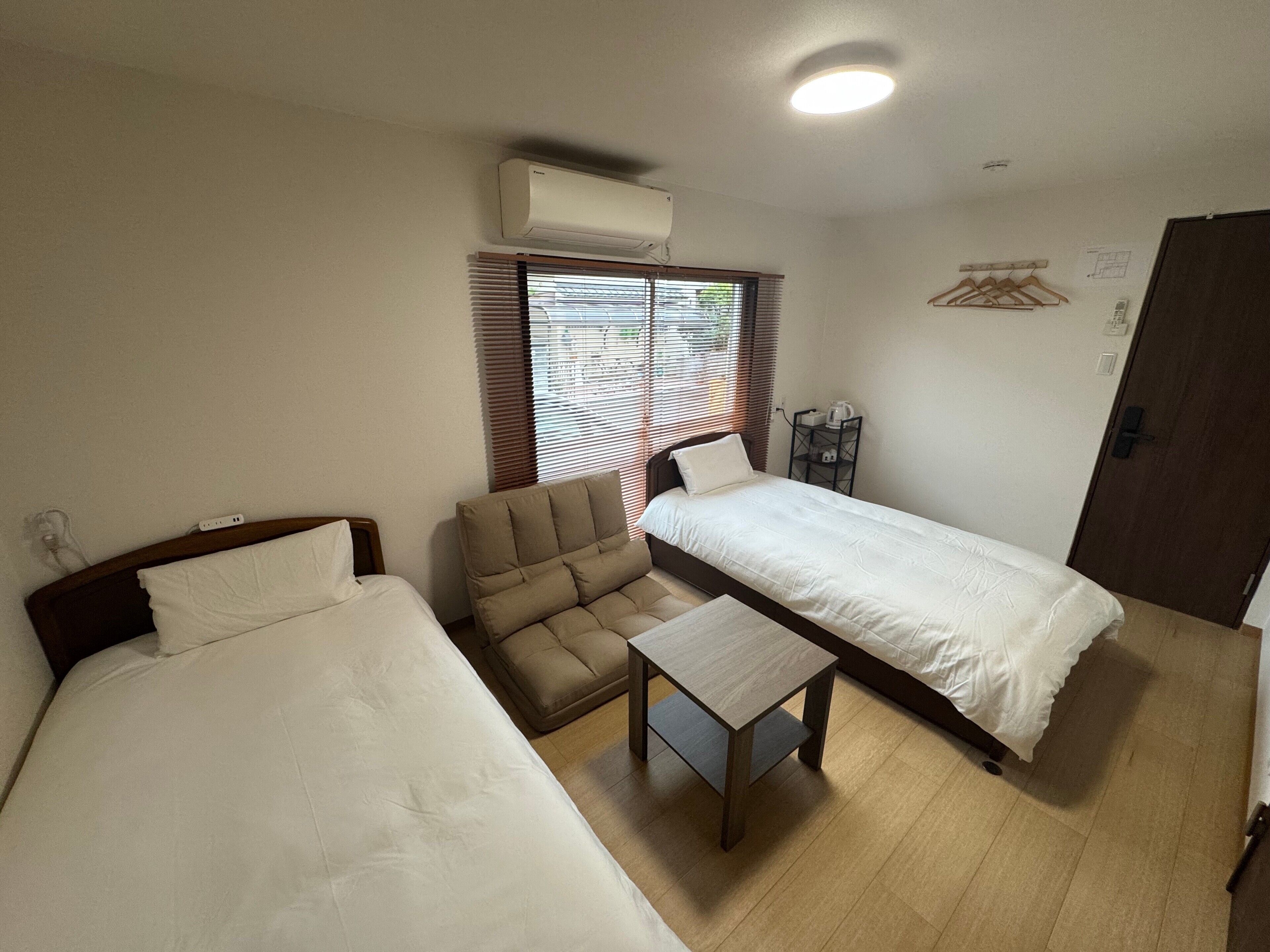 A room with two beds, a chair, a small table, an air conditioning unit, a window with blinds, a door, and a wall-mounted shelf with a kettle.