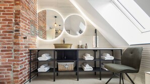 Un baño moderno con pared de ladrillo, un espejo grande, un lavamanos, repisas con toallas y una silla.