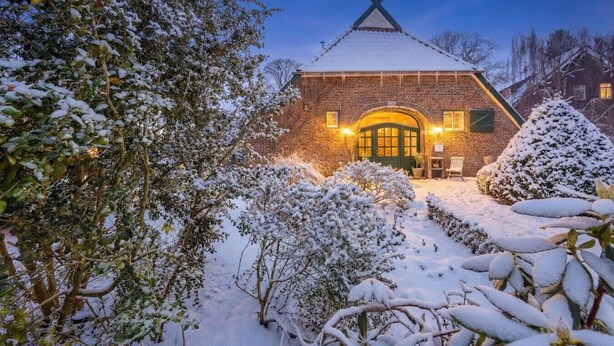 Un edificio de ladrillo con puerta verde y techo triangular, rodeado de árboles y arbustos cubiertos de nieve.