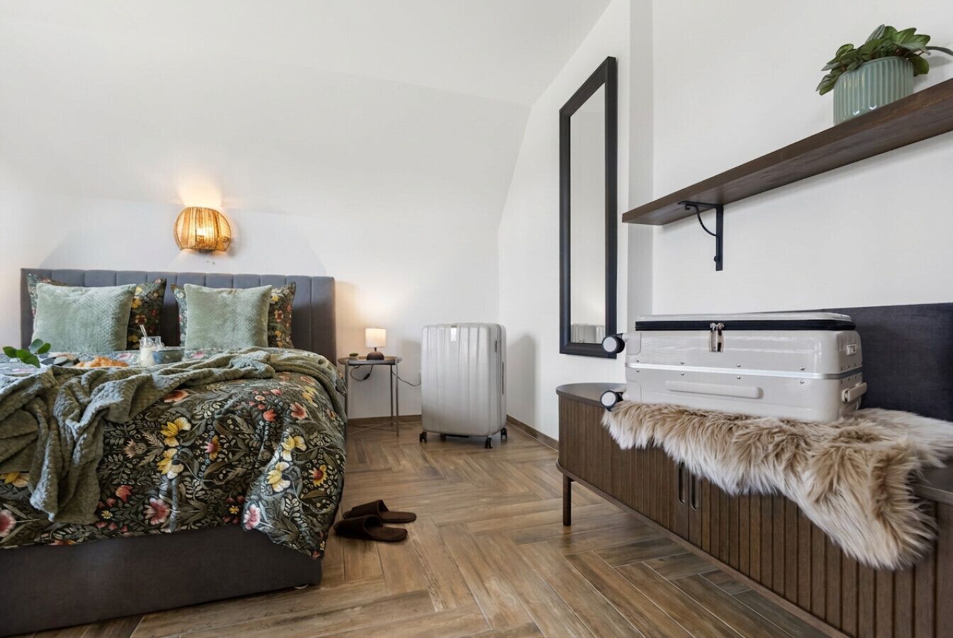 A bedroom with a bed, bedside table, lamp, suitcase, and a mirror on the wall.