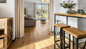 Una sala de estar moderna con un sofá, muebles de madera, un comedor con taburetes y vistas a través de un ventanal amplio.