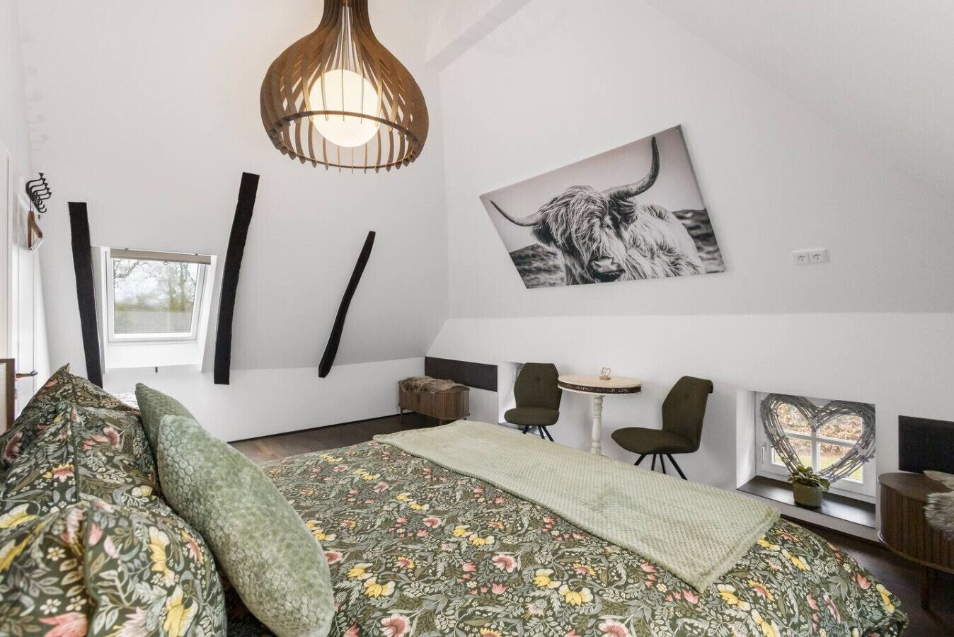 A bedroom with a bed, a desk, chairs, a hanging light fixture, and a bull's head artwork.