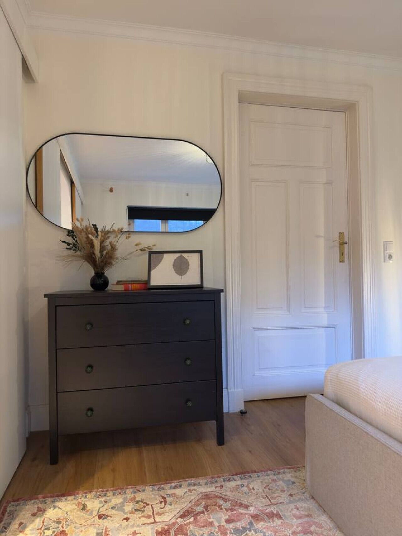 A room with a dark wooden dresser, a large mirror, a framed picture, a vase with dried plants, a patterned rug, and a white door.