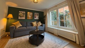 A living room with a sofa, a coffee table, a lamp, and a window with curtains. - The Forest Green Residence – Garden & Tranquility (Oldenburg)