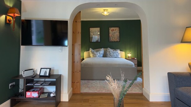 A bedroom with a bed, a TV mounted on the wall, and a console table with electronic devices. - The Forest Green Residence – Garden & Tranquility (Oldenburg)
