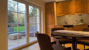 A kitchen with wooden cabinets, a dining table with chairs, and a view through large sliding glass doors to an outdoor seating area. - The Forest Green Residence – Garden & Tranquility (Oldenburg)
