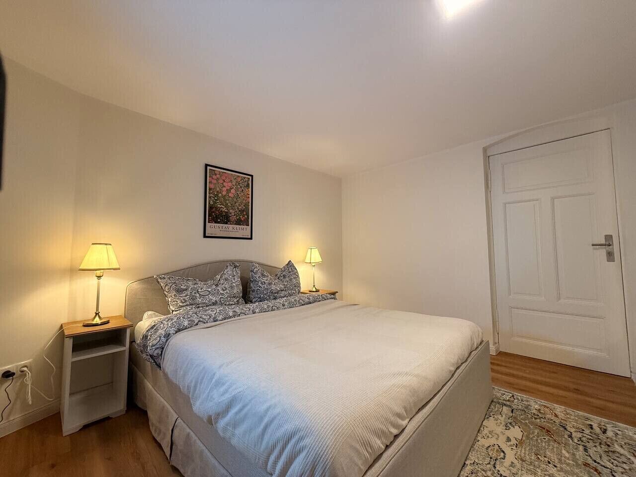 Quarto com uma cama, duas mesas de cabeceira com luminárias, uma porta e um quadro na parede.