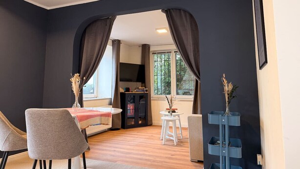 Une pièce comprenant une table à manger, des chaises, un téléviseur, une petite table avec un vase et une fenêtre avec des rideaux.