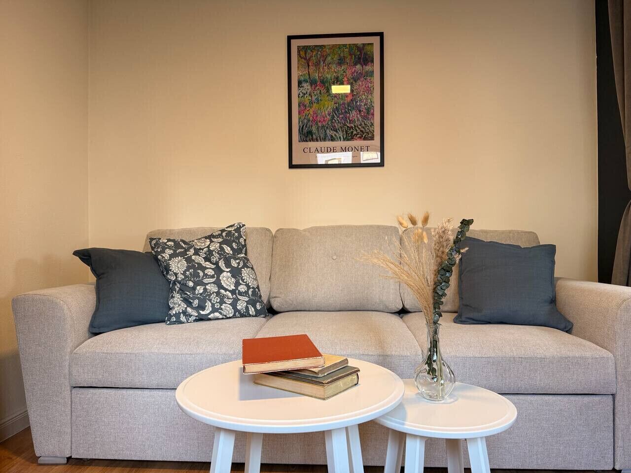 Een woonkamer met een grijze bank, twee witte bijzettafels, een lijstwerk boven de bank en een vaas met gedroogde planten op één tafel.