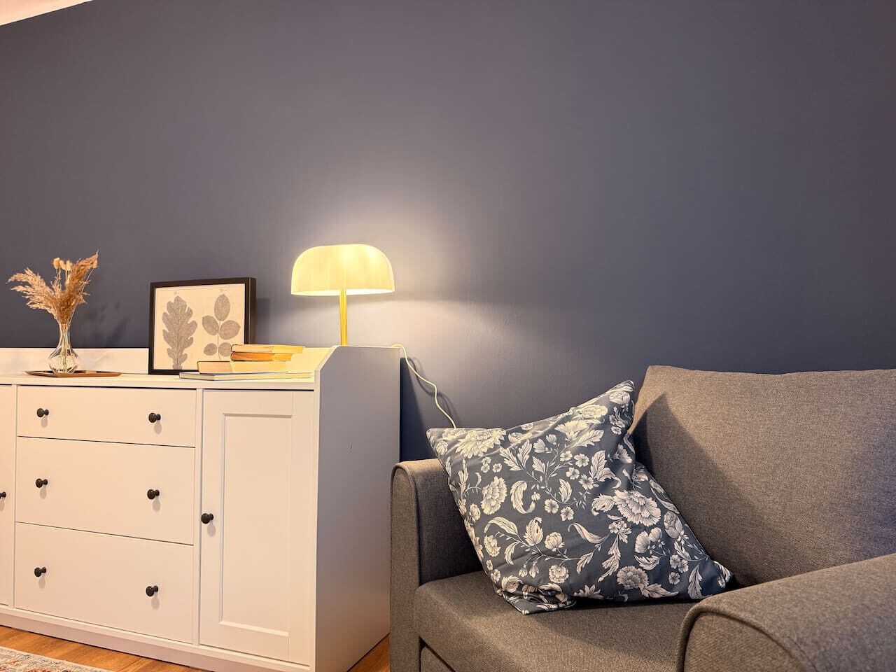 A room with a grey sofa, a white cabinet with drawers, a lamp, and a framed picture on the cabinet.