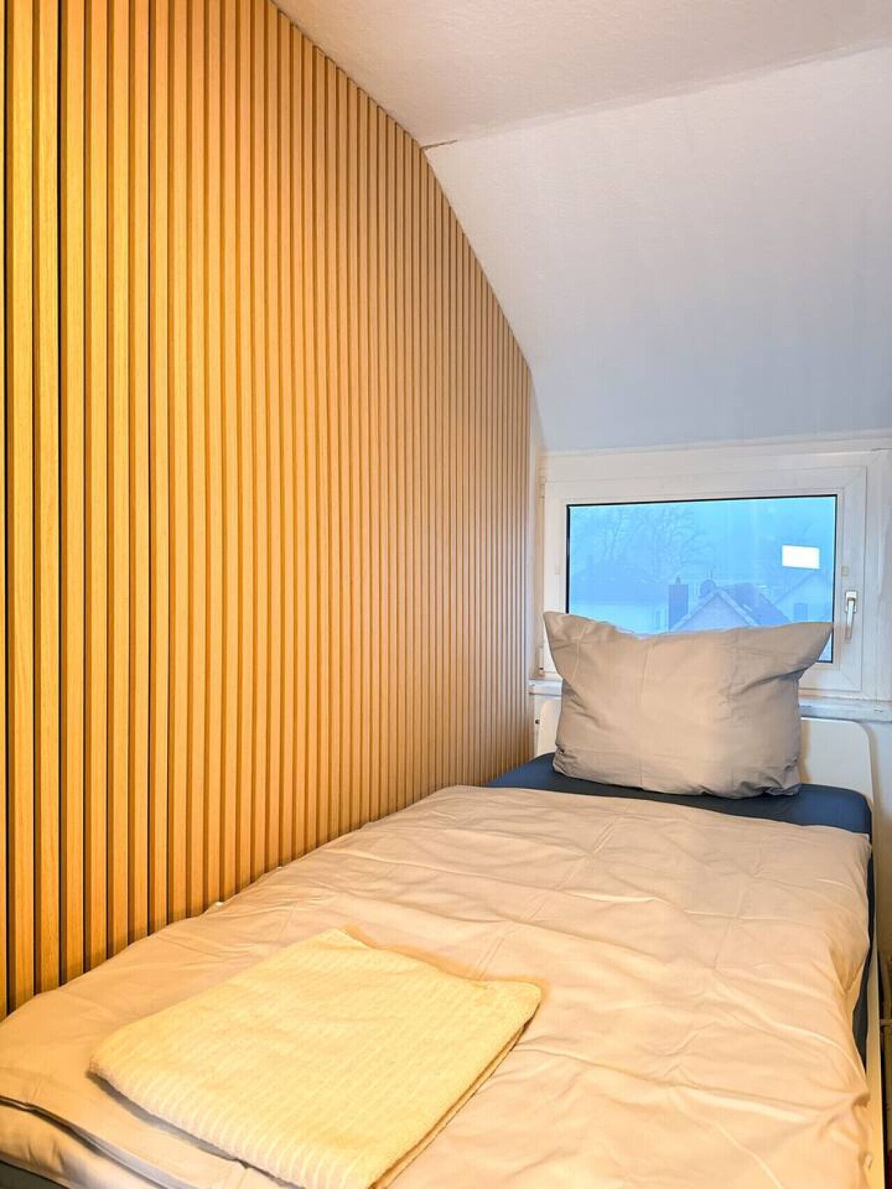 Ein Einzelbett mit beiger Matratze und Kissen, ein Fenster mit Blick auf Häuser und eine Wand mit Holzlatten.
