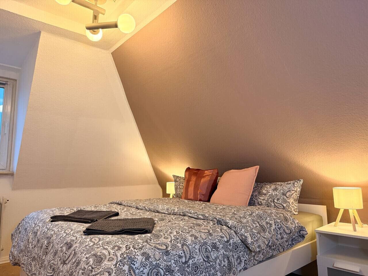 Un dormitorio con techo inclinado, una cama con ropa de cama estampada, dos lámparas de noche y una ventana a la izquierda.