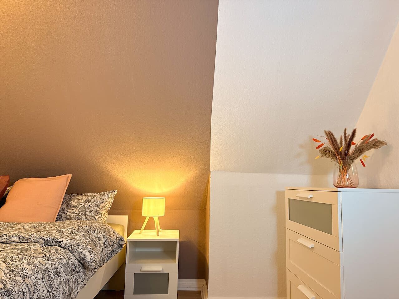Et soveværelse med en seng, et natbord med lampe, en kommode og en vase med tørrede planter.