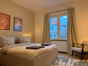 A bedroom with a bed, a chair, a window with curtains, and two framed pictures on the wall. - Peaceful 2BR Stay • Garden • Homey Comfort (Oldenburg)