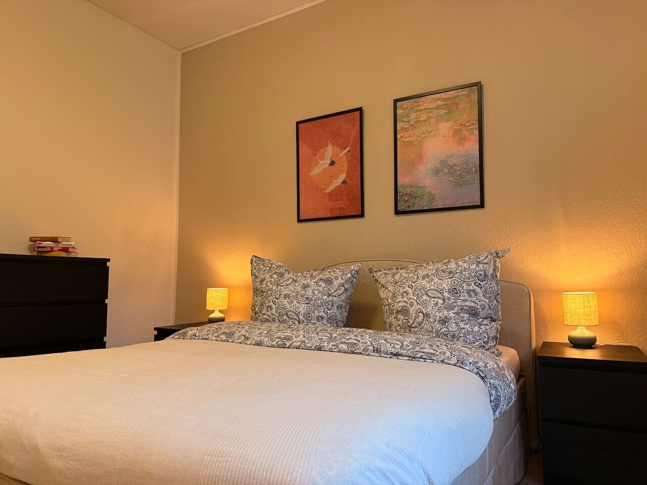 A bedroom with a bed, two bedside tables with lamps, a dresser, and two framed artworks on the wall.