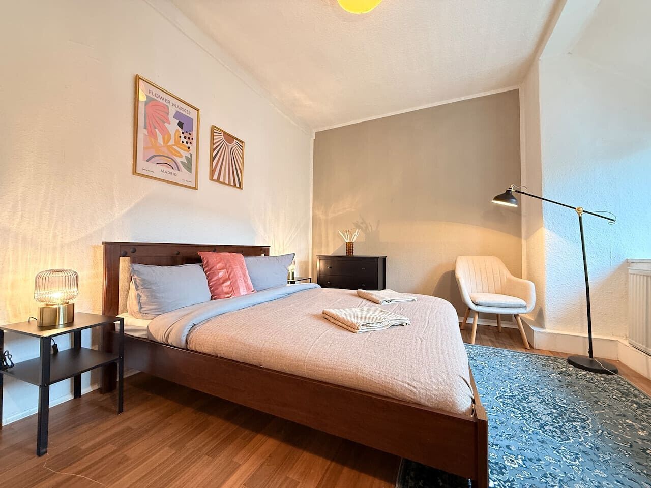 Une chambre à coucher avec un lit en bois, une table de chevet, une chaise, une lampe sur pied et deux œuvres d’art murales.