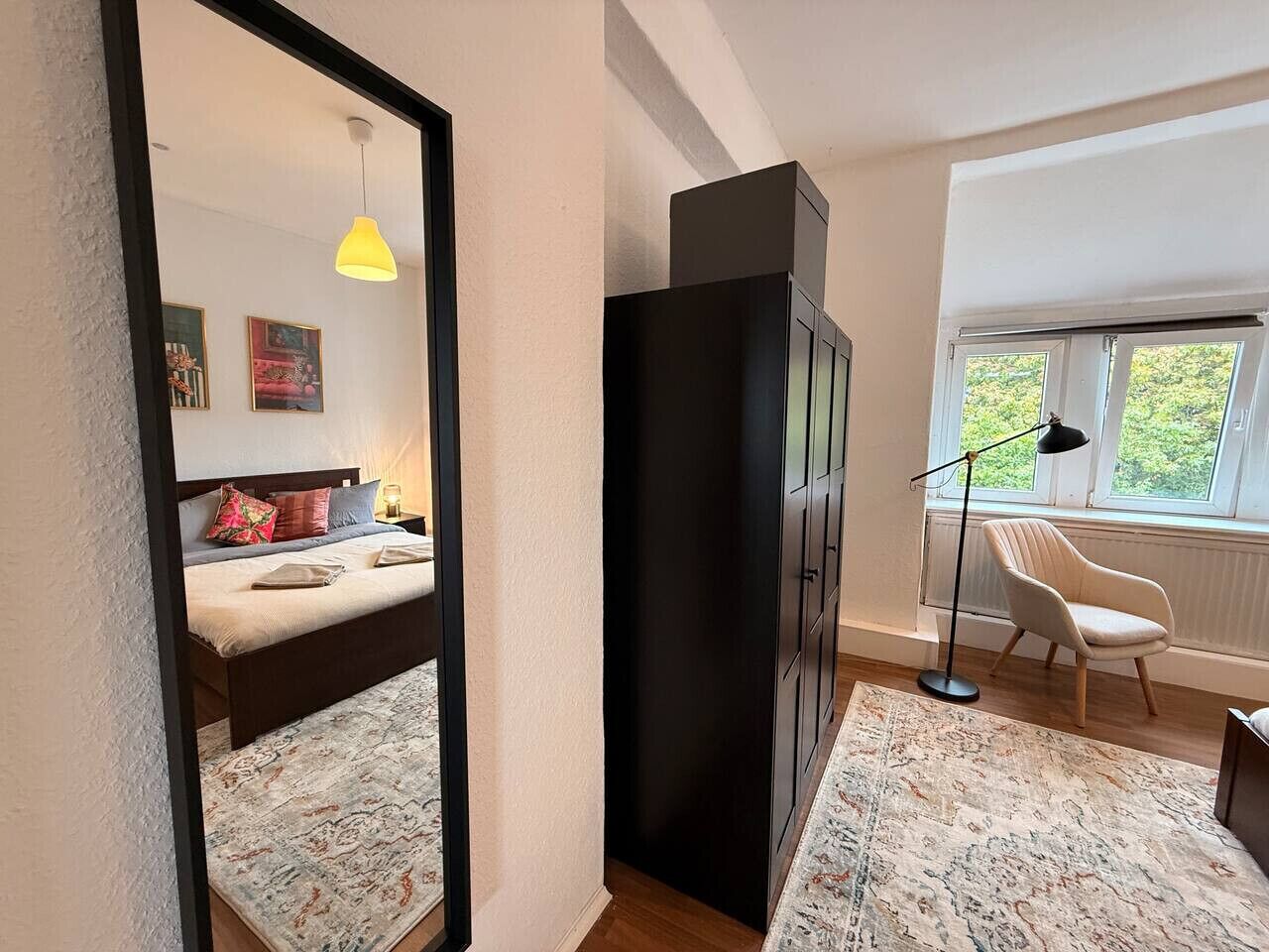 A bedroom with a bed, a wardrobe, a chair, a floor lamp, and a window with a view of greenery.