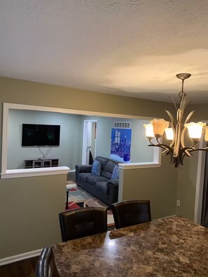 A dining area with a marble table, black chairs, a chandelier, a flat-screen TV, a gray sofa with patterned cushions, and a cityscape poster on the wall. - Cozy Family House 5 mins From Chiefs & Royals Stadium (Raytown)