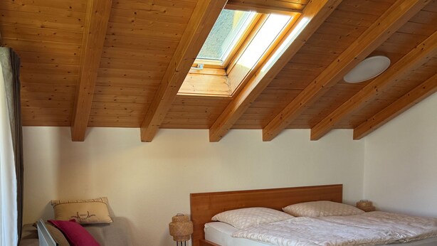 Une chambre avec un plafond en bois, un lit, une table de chevet, une chaise et une fenêtre.