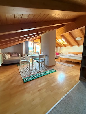 Ein gemütliches Dachzimmer mit Holzbalkendecke, einem Esstisch mit Stühlen, einer Couch und einem Bett.