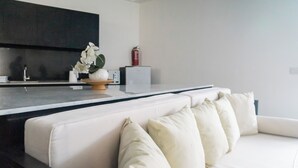 A modern living room with a white sofa, a kitchen area with dark cabinets, and a small plant on the counter. - Serenity Villa Nusa Dua (Badung)
