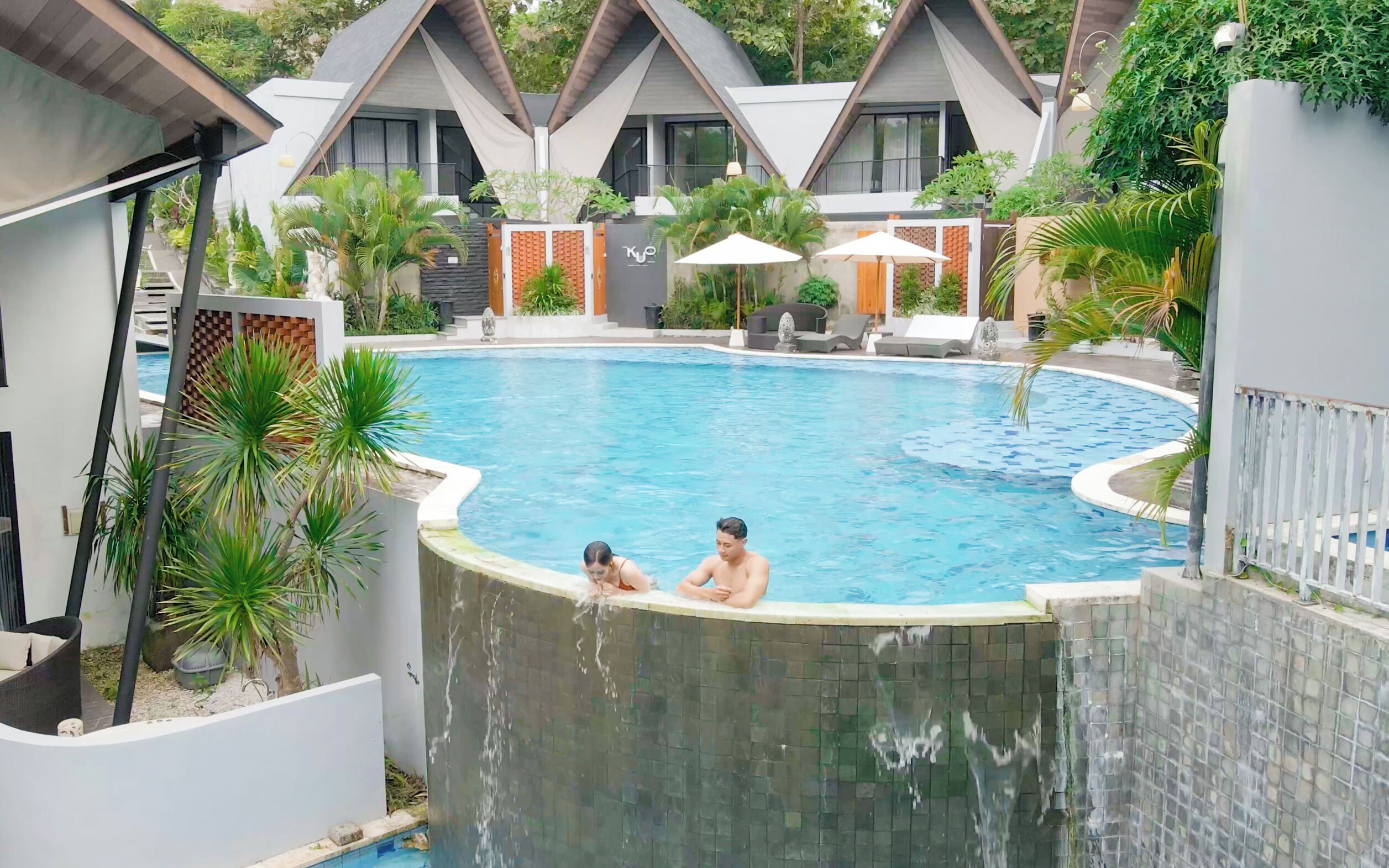 A swimming pool with two people sitting at the edge, surrounded by modern buildings and lush greenery.