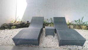 A set of three dark wicker lounge chairs with a small table, arranged on a pebbled surface with a stone wall and greenery in the background. - Serenity Villa Nusa Dua (Badung)