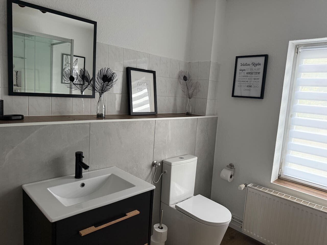 Une salle de bain avec un lavabo, des toilettes et un miroir. On y trouve des tableaux encadrés et une fenêtre avec des stores.