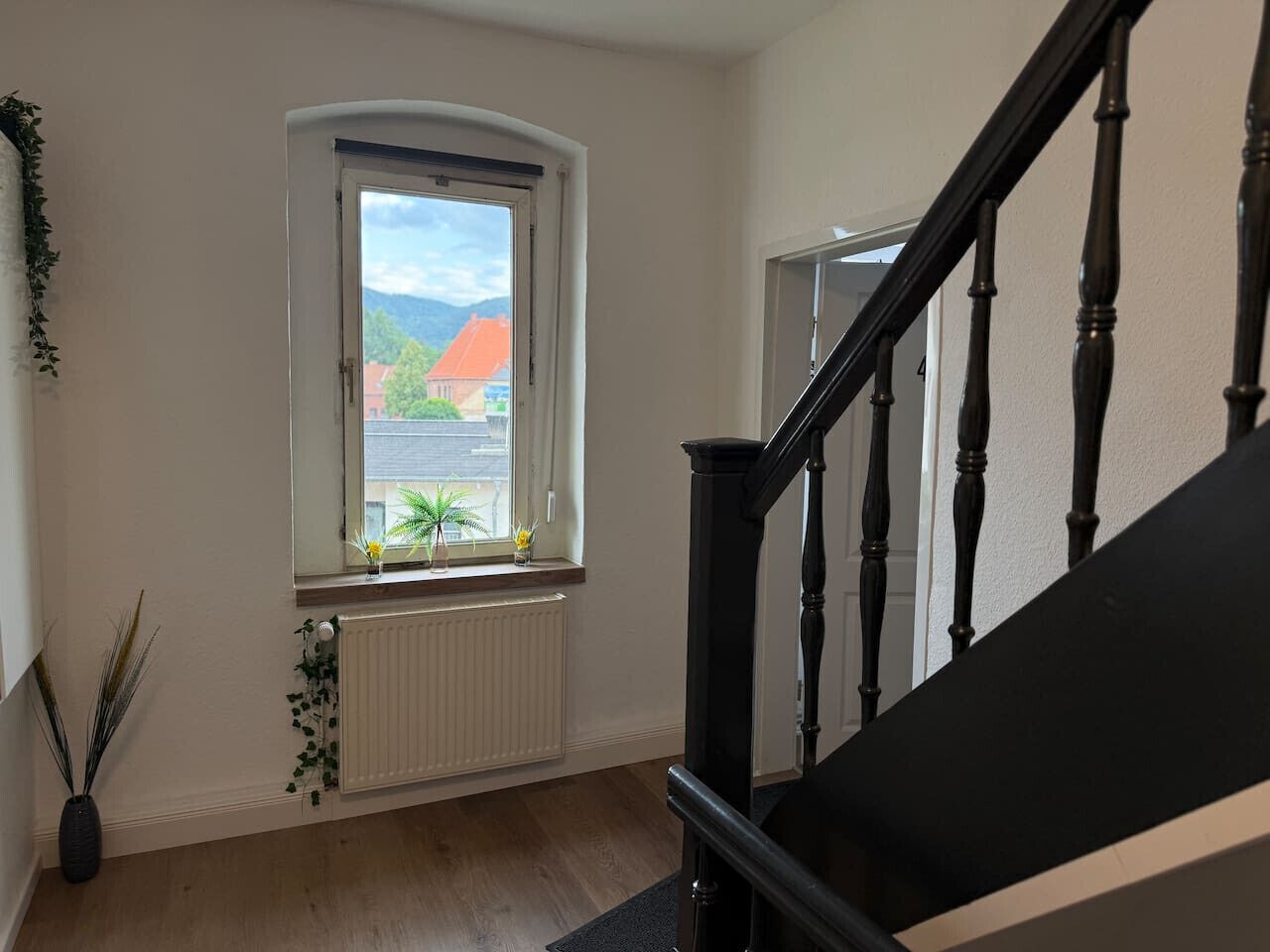 Eine Treppe mit schwarzen Spindeln, ein Fenster mit Blick auf ein Gebäude mit rotem Dach und ein Heizkörper unterhalb des Fensters.