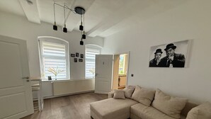 Un salon spacieux avec un grand canapé d’angle beige, une petite table basse en bois et un vase décoratif orné de plumes. Il y a deux fenêtres équipées de stores, une porte et une photographie en noir et blanc accrochée au mur.