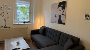 Una sala de estar con sofá, mesa de centro, lámpara, ventana con vistas y una obra de arte en la pared.