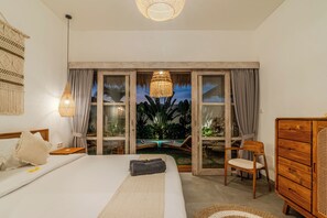 Ein Schlafzimmer mit einem Bett, einer Holzkommode, einem Sessel, einem Fenster mit Vorhängen sowie Blick auf einen Pool und Grünflächen.