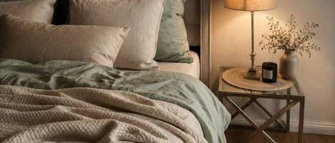 A bed with a green and beige blanket, pillows, and a bedside table with a lamp and a vase.