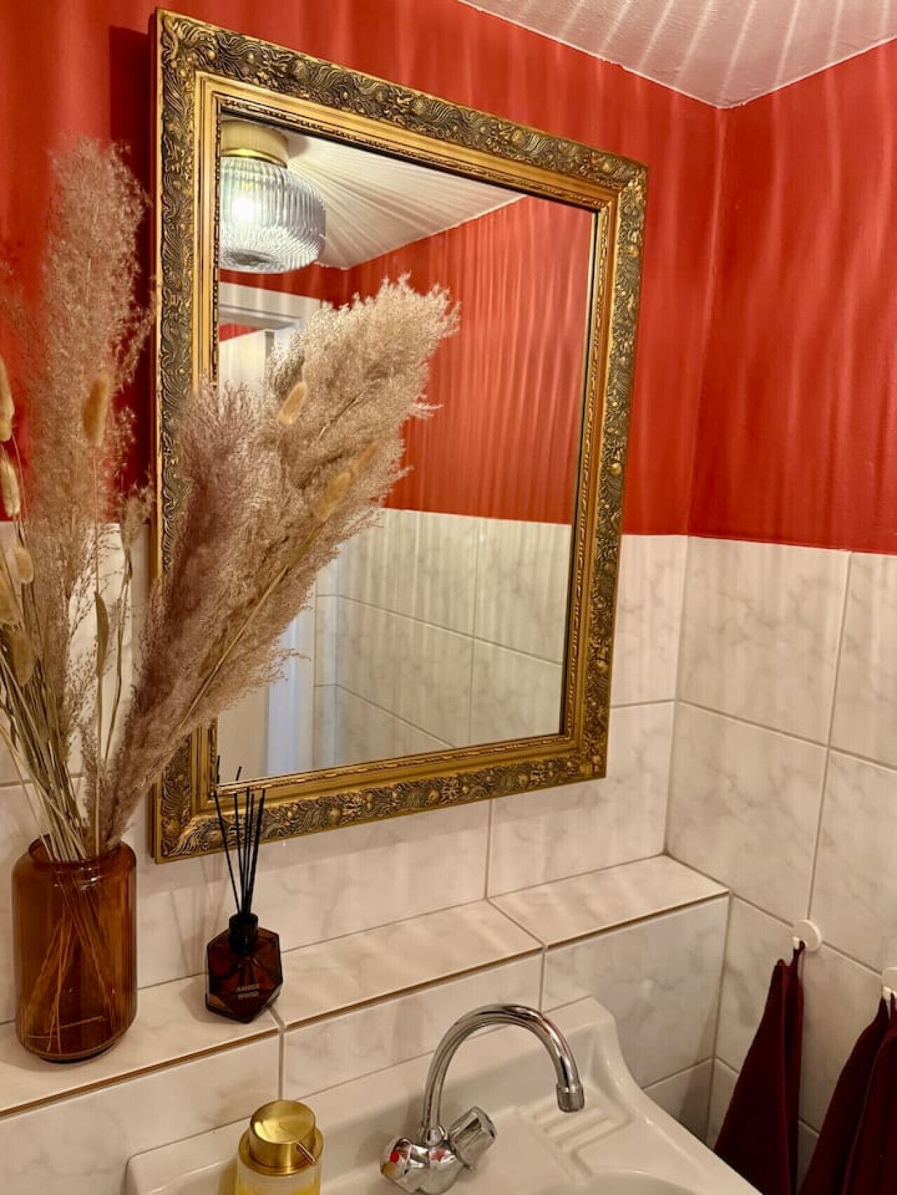 A bathroom with a large ornate mirror, a vase with dried plants, a bottle of hand soap, and a towel rack.