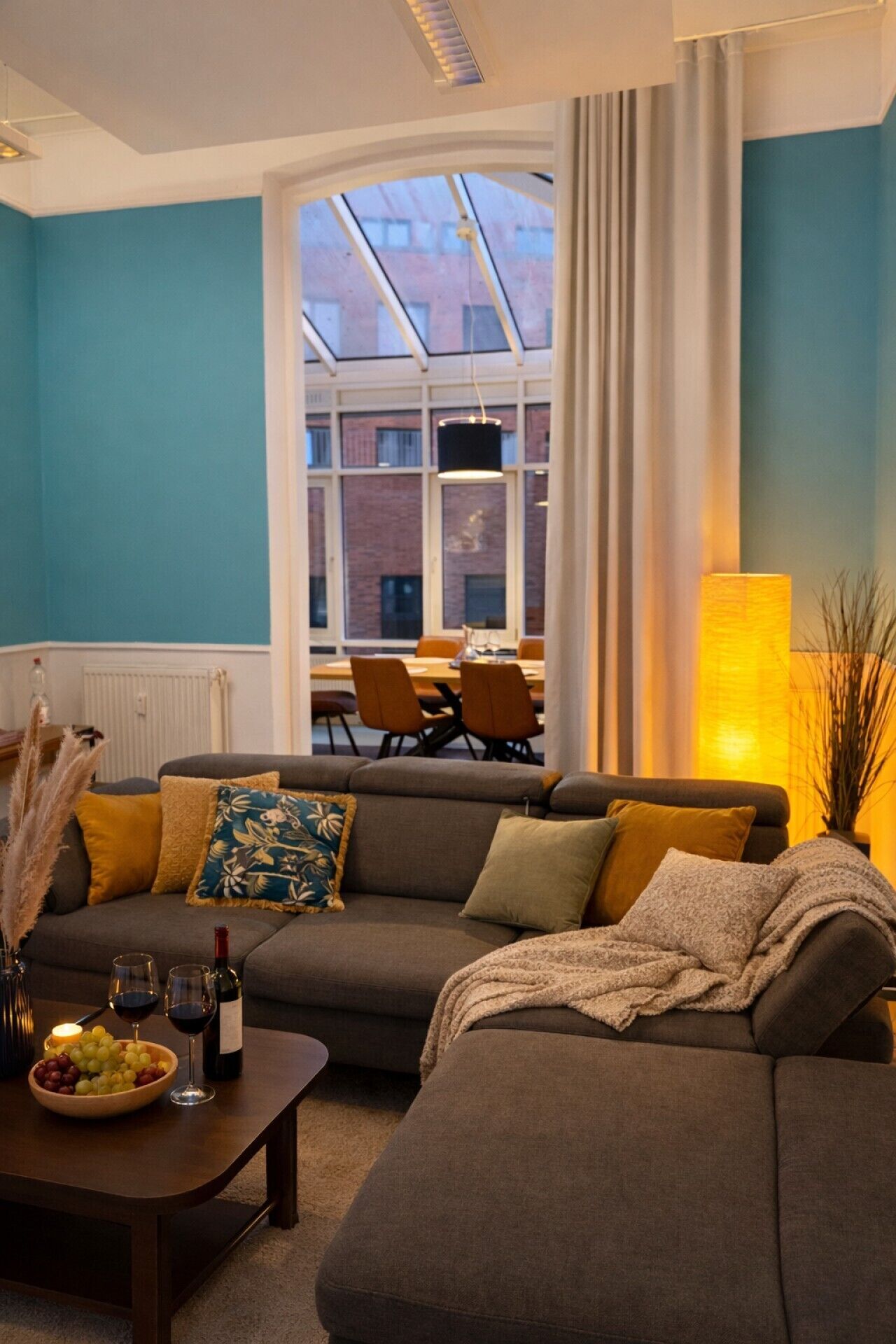 A modern living room with a sofa, a coffee table with wine glasses and grapes, and a view of a dining area through large windows.