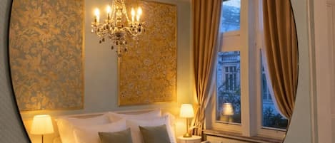 A hotel room with a bed, a chandelier, a window with curtains, and a wall with floral wallpaper.