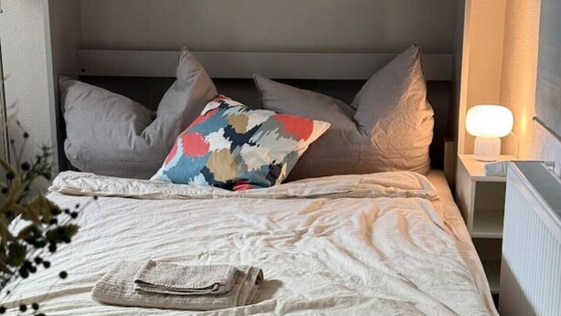 Una cama bien hecha con almohadas, una mesita de noche con una lámpara, y un plato con un vaso sobre una mesa.