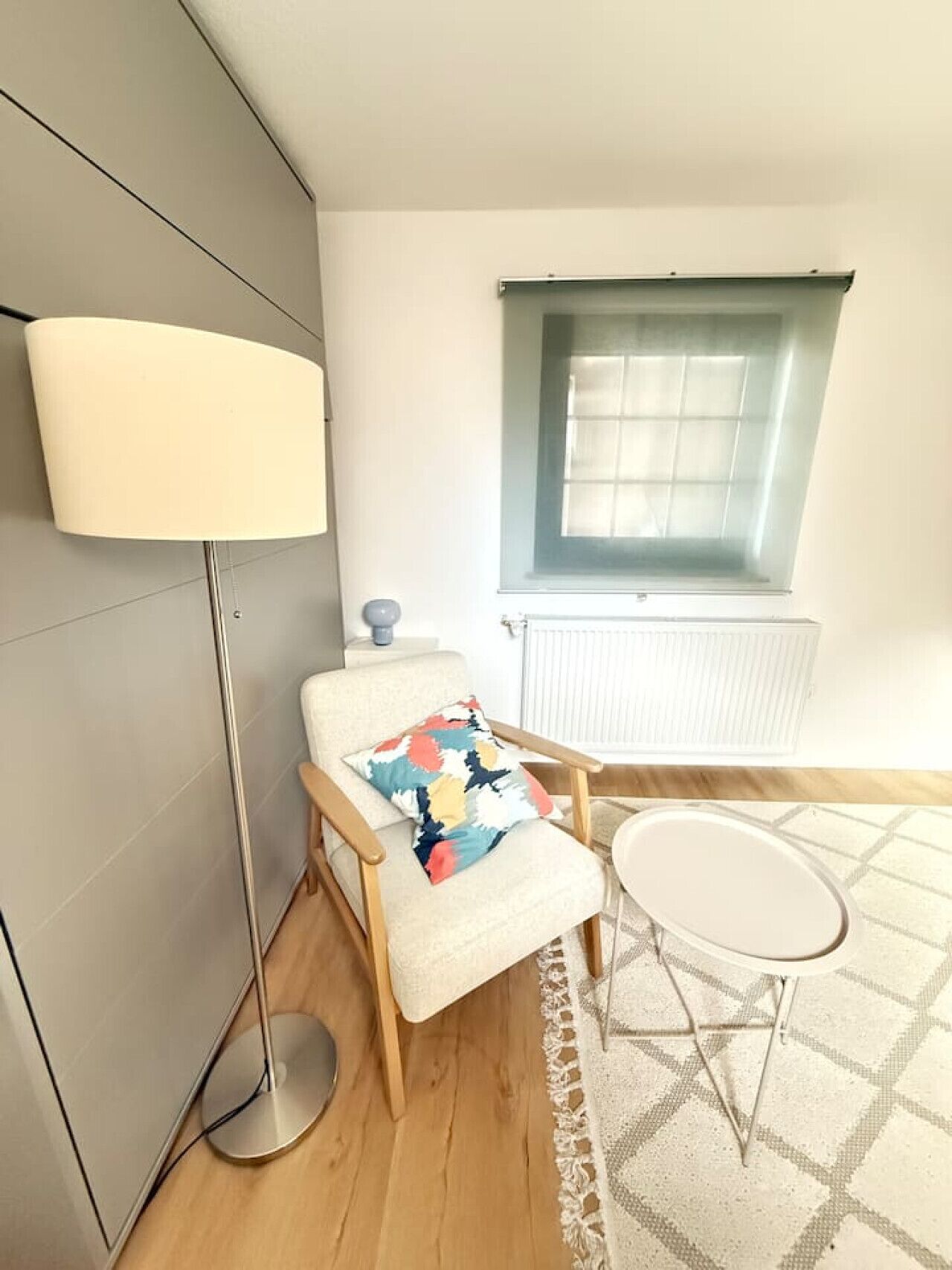 Ein modernes Zimmer mit einer Stehlampe, einem Sessel mit farbenfroher Sitzkissen, einem kleinen Rundtisch und einem Fenster mit Jalousien.