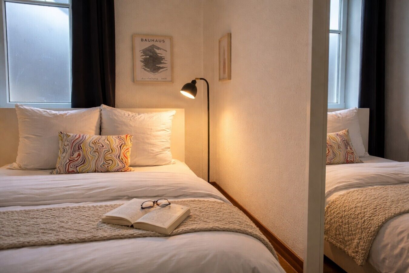Ein Hotelzimmer mit zwei Betten, einem gerahmten Bild an der Wand, einer Nachttischlampe und einem Buch auf dem Bett.