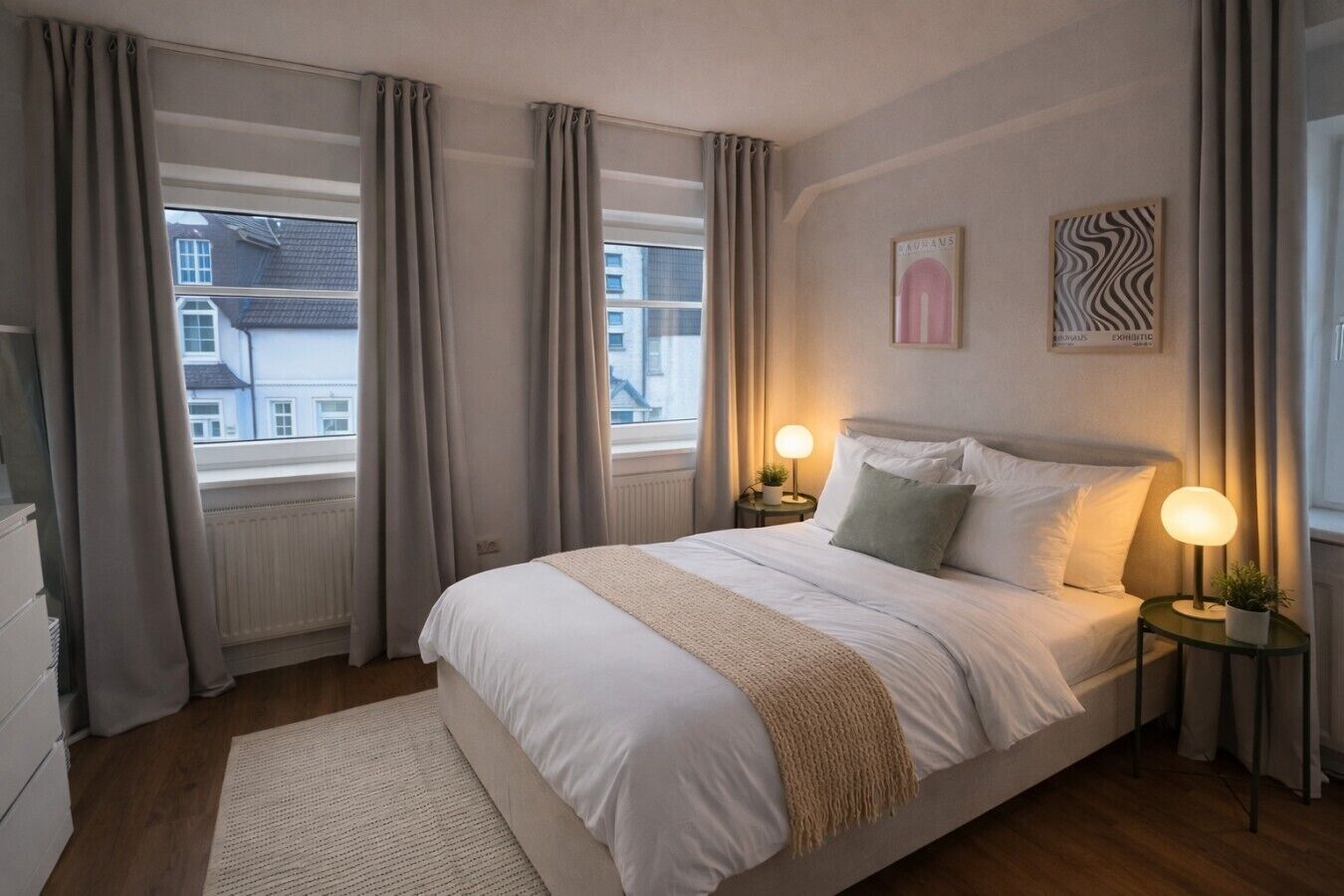 A bedroom with a large bed, two bedside lamps, a window with curtains, a radiator, and a small plant on the bedside table.