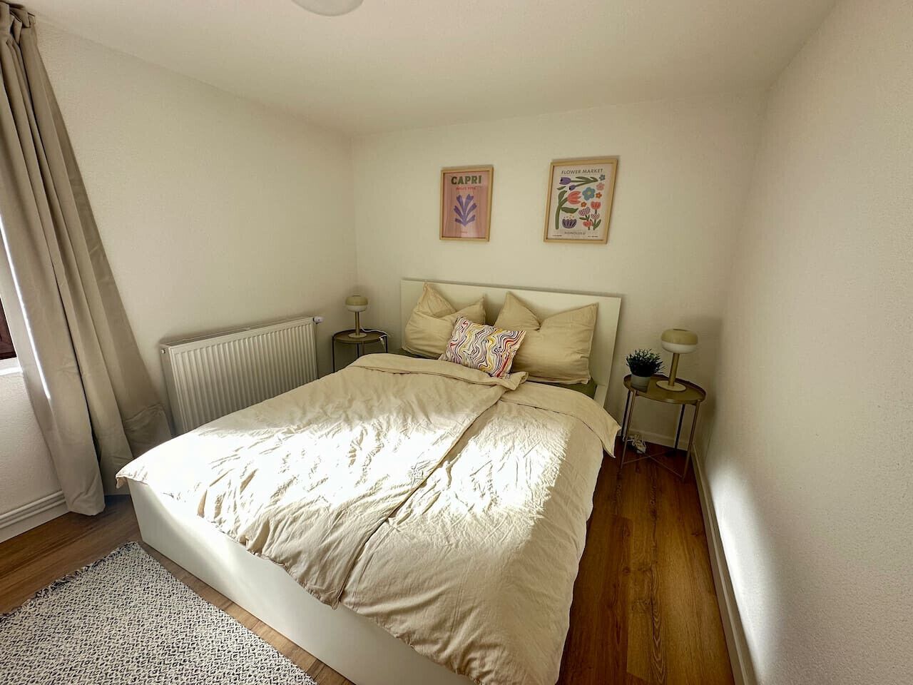 Una camera da letto con un letto, due comodini, un termosifone, una finestra con tende e due quadri appesi al muro.