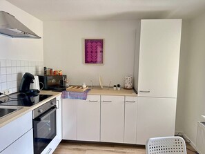 Eine kleine Küche mit weißen Schränken, einem schwarzen Herd, einer Kaffeemaschine und einem Kühlschrank.