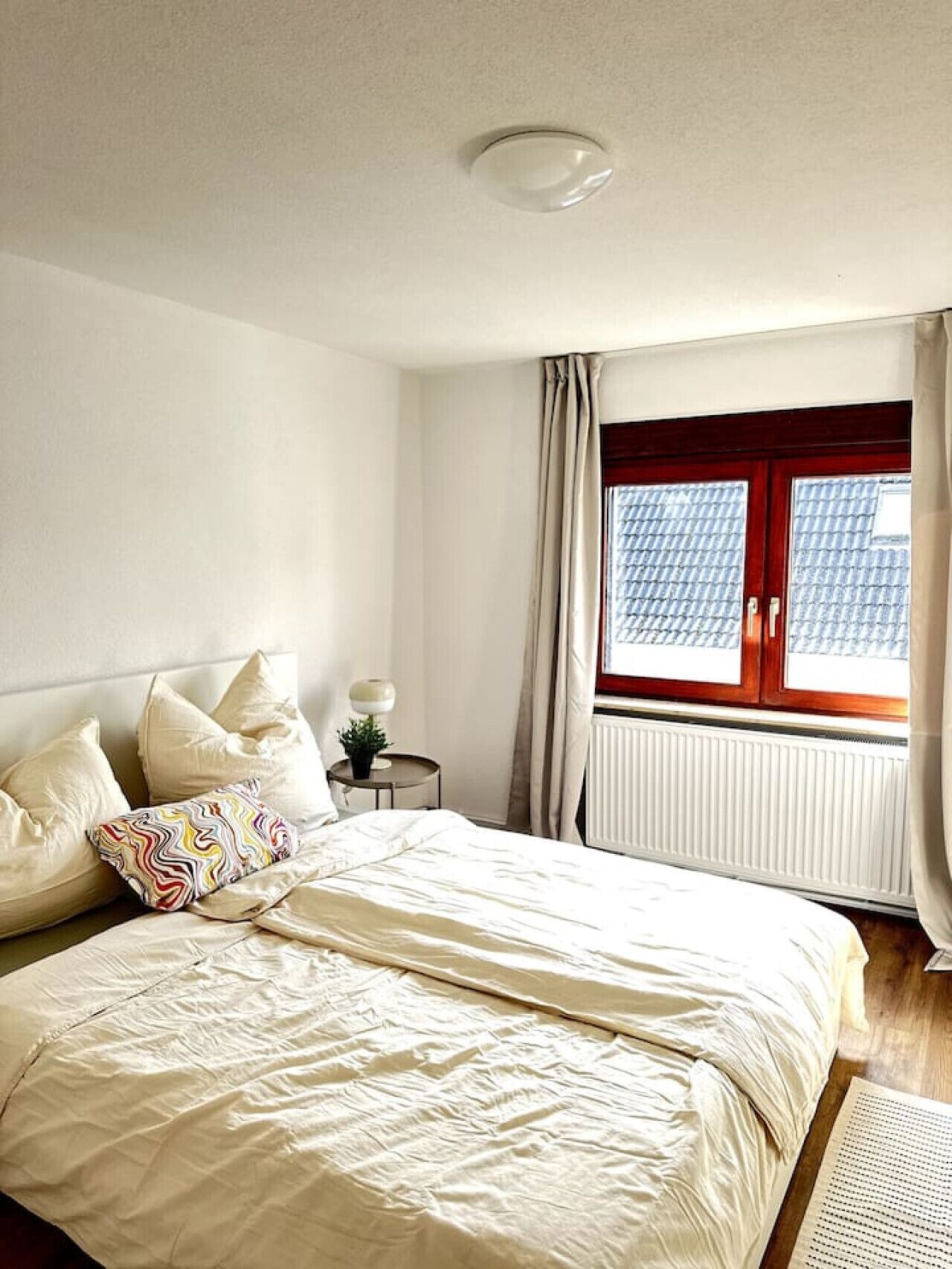 Ein Schlafzimmer mit Bett, Kissen, Nachttisch mit Lampe, Fenster mit Vorhängen und Heizkörper.
