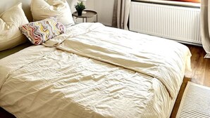 Une chambre à coucher avec un lit, des oreillers, une table de chevet avec une lampe, une fenêtre avec des rideaux et un radiateur.