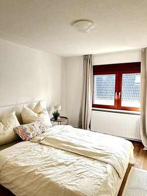 Ein Schlafzimmer mit Bett, Kissen, Nachttisch mit Lampe, Fenster mit Vorhängen und Heizkörper.