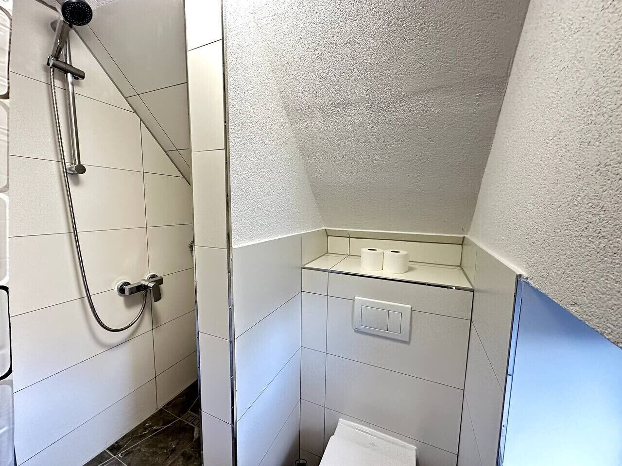 Ein kleines Badezimmer mit weiß gefliesten Wänden, einer Dusche mit Handbrause, einer Glasablage mit Toilettenpapier und einem Handtuchhalter.