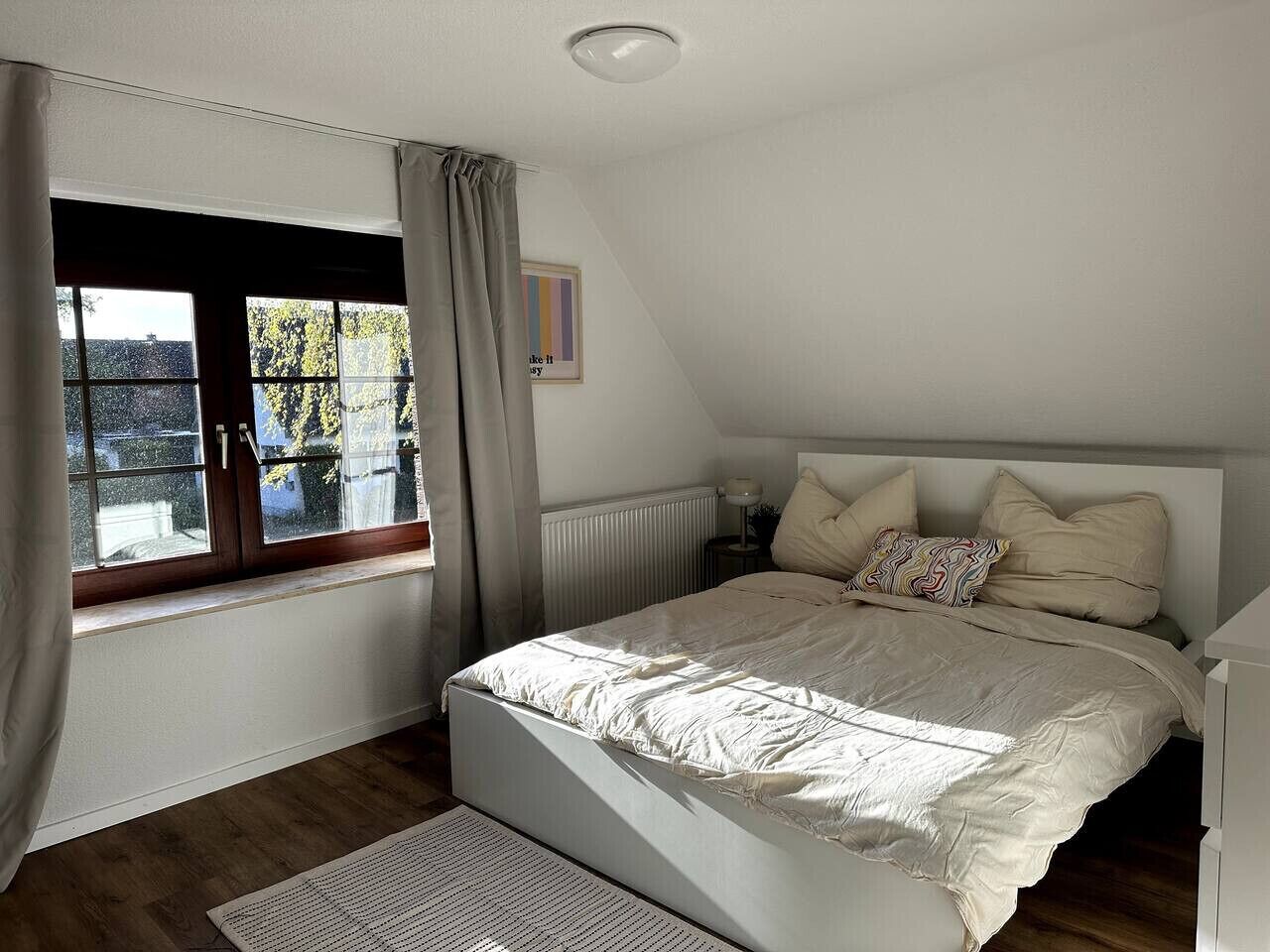 A bedroom with a bed, a window with curtains, a radiator, and a small table with a lamp.
