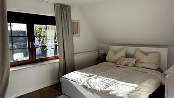 A bedroom with a bed, a window with curtains, a radiator, and a small table with a lamp.